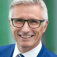 A smiling man with gray hair, wearing glasses and a blue suit, poses for a photo against a blurred green background.