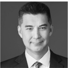A black and white headshot of a person wearing a suit and tie against a plain background.