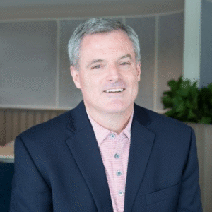 Man in a dark blazer and striped shirt smiling indoors with a plant in the background.